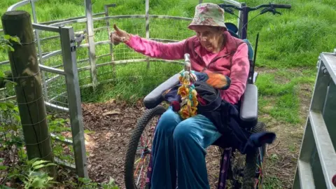An elderly woman in a wheelchair gives a thumbs up as she goes through a gate on a rural path. she is wearing a tan patterned summer hat, a pink jacket and blue trousers.