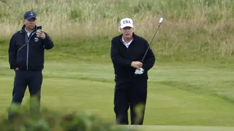EPA Trump completes his golf swing and follows looks as his ball heads off. A man in golfwear and hat and filming on a phone is behind him