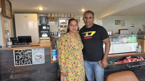Martin & Jones Marketing A man and a woman are standing inside a cafe in front of a dark wood counter with a screen and small black receipt printer on it. There is a glass fronted container on it and there are pastries inside it