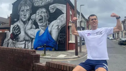 Joe Wild Joe Wild during his marathon in Liverpool, posing with his biceps flexed in front of a mural of martial arts fighters Paddy Pimblett and Molly McCann.