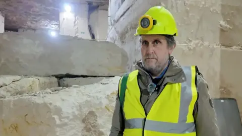 Seb Brookes is wearing a yellow hi-vis jacket and hardhat with flashlight attached. Beneath that he's wearing a heavy green coat. Behind him is a pile of white quarried stone. 