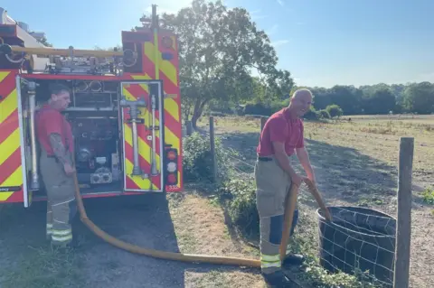 London Fire Brigade Two male firefighters in uniform standing behind a fire truck in a field. The man on the right is smiling, looking directly at the camera as he holds a hose inside a bucket which is on the other side of a small fence that sits just above his knees. The hose is connected to the back of the fire truck. They are wearing red T-shirts, khaki trousers with reflective stripes near the end and black boots.