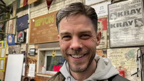 A man with brown hair and a beard stands smiling at the camera in a boxing club. He is wearing a grey hooded sweatshirt. There are posters on the wall behind him with words like Kray, amateur boxing and Repton. There is also a small booth-style window with a red digital clock in front of the glass. 