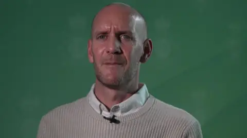 BBC Dan Barr, 41, who has blue eyes, a bald head and light stubble, speaks into the camera wearing a beige jumper over a white shirt.