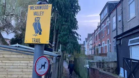 Elizabeth Baines / BBC A sign which reads Meanwood Valley Trail hangs above an alleyway. A row of terraced houses line the other side. The sign is yellow with a black owl illustration