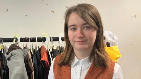 A woman in a white shirt and an orange waistcoat stands in front of a clothing rail with lots of clothing hanging on it.