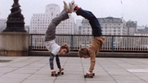 Two teenagers doing handstands on their skateboards, with their feet touching in the air.