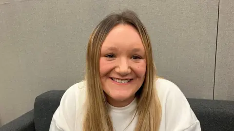 A close-up image shows a person seated on a dark chair against a plain textured backdrop. The person has long, light-coloured hair and is wearing a light-coloured top, facing directly toward the camera.