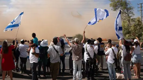 AFP via Getty Images Dozens of people stand on the side of the road. They were waving Israeli flags and in front of them was a large cloud of dust.