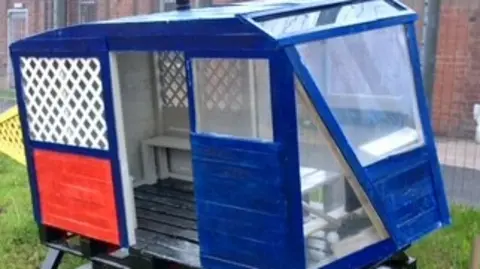 OPAL A blue and red bus-looking item made out of ballets. It has windows and a door with white seats inside. It is on a black base, sitting on a grass verge. Around it is metal fencing and a brick building behind. 