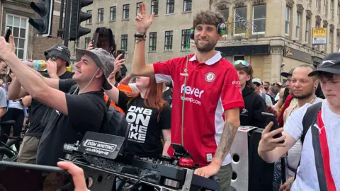 Dom is on his bike with lots of people around him filming on their phones. He is wearing a white and red Bristol top and has his hand in the air.