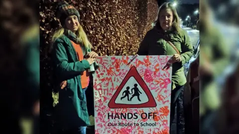 Jenny Holmes and Bridget Petty stand in the dark, wearing coats and Jenny wearing a woolly hat. Between them they are holding a poster with red painted handprints covering it, and a children walking road sign drawn into the middle. Under the sign the slogan reads - 'HANDS OFF Our Route to School'