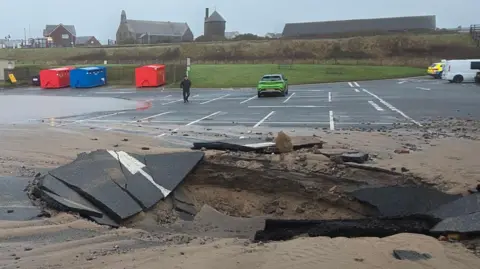 Shane Stanbridge The sinkhole in a tarmac car park. The hole is about as large as three parking spaces. The area surrounding the hole has broken sections of tarmac and sand around it. One side of the car park is covered in water. A handful of cars are parked some distance away from the hole.