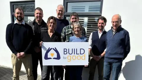 Build for Good Rachel Rogers and Sarah Thornton stood in the middle of a group of people holding a white sign which says "Build for good" and the logo, on the left, is two blue hands making the shape of a house with a yellow heart in the middle. Around them is six men. The building behind them is white with two windows. 