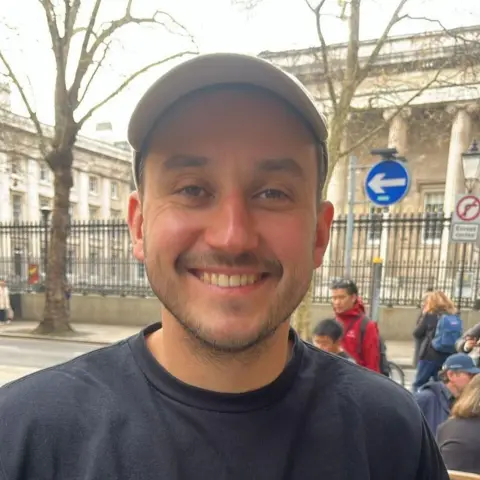 Selfie of young man in navy T-shirt, wearing a beige baseball cap. He's smiling at the camera. 