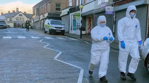 Julie Smith/BBC Two masked investigators are walking down a main road wearing full white body-suits and blue protective gloves. A police officer and van can be seen behind them further up the road. Police tape is on a post at a zebra crossing.