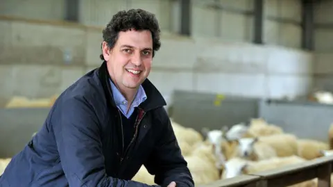 Professor David Main Professor David Main wearing blue overalls and smiling at the camera with sheep behind him. 