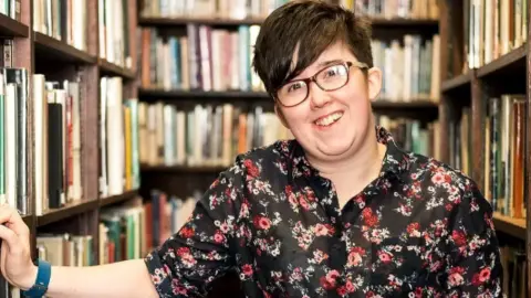 Brendan Gallagher Lyra McKee is wearing a black and red floral shirt.
She is standing in front of bookcases in a library.
Her hand is rested on a shelf and she is smiling.