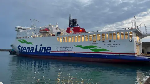 The Stena Vinga ferry is pictured in St Helier marina in Jersey. It is a white vessel and a red lifeboat is attached to the side. The words Stena Line Connecting Europe for a Sustainable Future are written on the side in blue. 