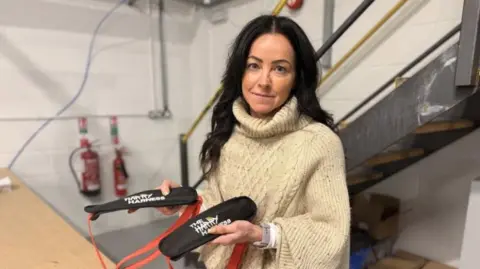 Joanne, who is wearing a cream knitted top and jeans, is holding the harness which has orange straps and a black pad with the words The Harry Harness in white writing. 