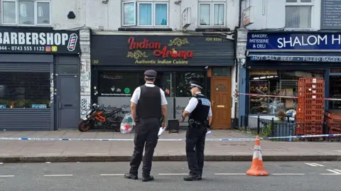 PA Media Two police officers in uniform stand on a road in front of police tape which surrounds shops and restaurants. Their backs are to the camera. Signage for the businesses pictured indicate they are a barbershop, an Indian restaurant and a deli.