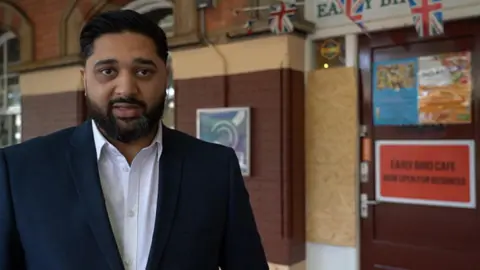 A man in a white shirt and blue suit jacket stands in front of a cafe at a railway station, which has a boarded up window.