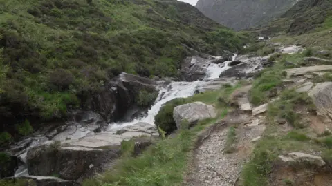 BBC A stream cascades down a series of rocks alongside a walking path. The bank slopes upwards on the left hand side.