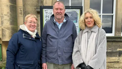 Julia Bryson/BBC A woman with short blonde hair and a navy coat, a man in a blue jacket and grey trousers and a woman with blonde curly hair and a long grey fur-style coat, stand outside a civic building in Ilkley. 