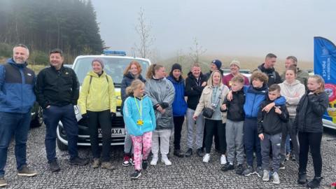 A group of adults and children pose in front of police cars parked in a grey and rainy hilly landscape in Wales