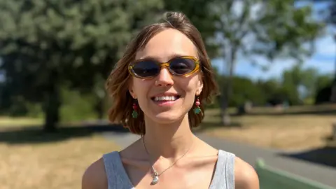 Robyn Surcombe - a young woman with brown hair, wearing sunglasses and a grey vest top, smiles at the camera