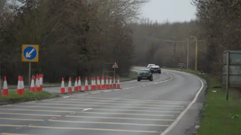 BBC A single carriageway stretch of road showing traffic cones and a keep left sign ahead of a bend to the left. Three cars can be seen in the distance in the left lane heading out of shot.