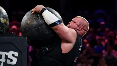 Kane Francis competing in Giants Live strongman competition. He is lifting an atlas stone on to a platform that is head height. The smooth spherical stone is so large he cannot get his arms around it. His face is red with exertion.