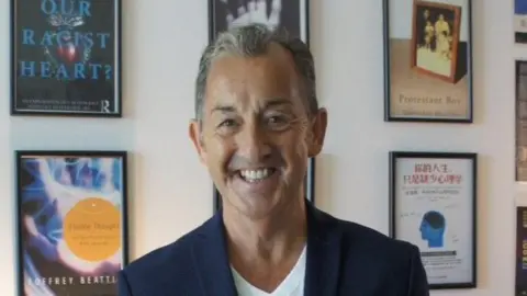 Geoff Beattie Prof Geoff Beattie smiles in front a wall showing framed pictures of some of his books. He wears a dark suit jacket over a white top.