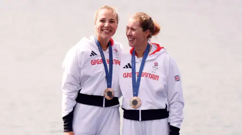 PA Media Rebecca Wilde and Mathilda Hodgkins-Byrne. They are both blonde and pictured wearing white Adidas jackets with Great Britain printed across the chest in red letters. They both have their medals around their necks and are smiling. 