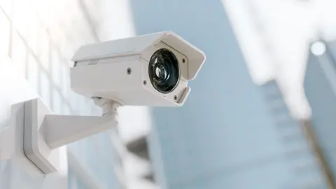A CCTV camera on a wall with blurred buildings in the background