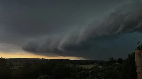 Weather Watchers/Robin Thomas A landscape scene with fields and trees below and a very dark sky with a band of very storm-like clouds swirling into the distance. There is a light glimmer of sunshine low on the horizon