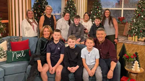ITV Three boys from the Denny Boys sitting on a sofa on the This Morning set between hosts Cat Deeley and Ben Shepherd. Behind the couch, two other boys are standing next to their mums.