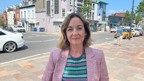 A woman with a balayaged long bob, wearing a pink jacket, striped top stands on a sunny day on Holywood High street staring at the camera.
