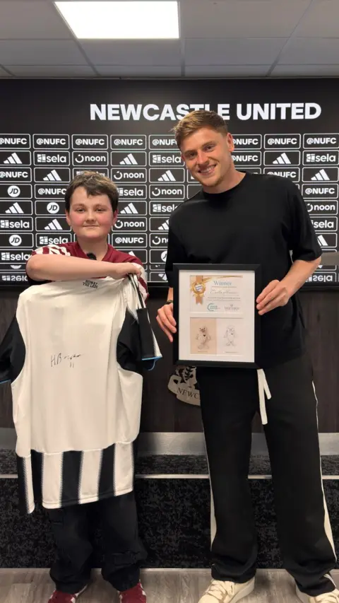 Young boy stands with signed Newcastle United shirt and player Harvey Barnes.