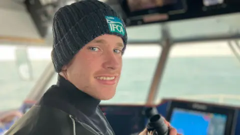 Fiona Irving / BBC A white man smiles at the camera. He has a black beanie-hat on with the IFCA blue logo on it. He is in holding some binoculars - out of the windows around him is water. There is a computer screen in front of him.