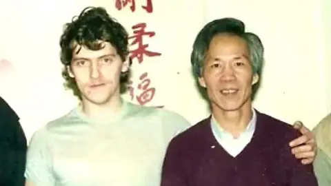 Liverpool Hung Gar Kung Fu School Stephen Ornellas (left) in an image from the 1980s with brown wavy hair wearing a pale green t-shirt has his arm around Master Jimmy Chan who has greying black hair wearing a white shirt and a burgundy jumper. There is Chinese writing in red on the white wall behind them. They are slightly smiling. 