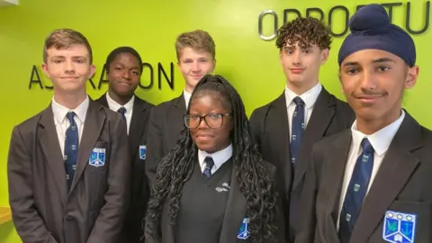 Alex Pope/BBC Six pupils at Wootton Upper School wearing school uniform standing in front of a green wall. All six are smiling at the camera and they are all wearing blazers and blue ties