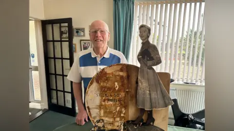 A smiling Reginald Wright looks directly at the camera as he stands in his living room behind the sign. The sign has a woman in high heels and a midi-length dress standing and carrying a clutch handbag. 