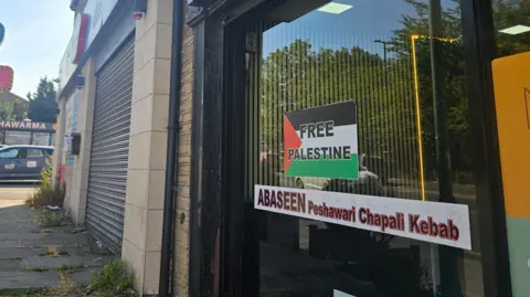 Aisha Iqbal/BBC A shop window in a street with several businesses. Signage identifies it as a kebab shop. A small poster in the window features a black, white, red and green flag with the words 'Free Palestine' on it.