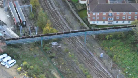 City of York Council An aerial photograph of the existing Wilton Rise footbridge crossing railway lines in York. The bridge runs horizontally across the centre of the image, spanning several railway tracks below. On the left there is a rail yard containing two stationary rail vehicles. A cluster of white vans is parked in a yard in the bottom-left corner.