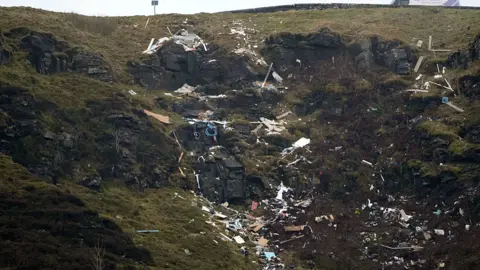 Waste scattered down a mountain with a lay-by above. It's not possible to pick out individual items but there are sheets of wood and small items covering the rocky ground.