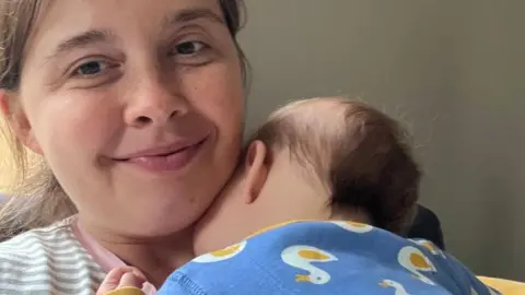 Jessica Kent-Hazledine cuddles her baby son, whose face is next to her neck. Kent-Hazledine is smiling. She is wearing a green and white stripey t-shirt. Her son is wearing a blue babygrow with white and yellow ducks on it.