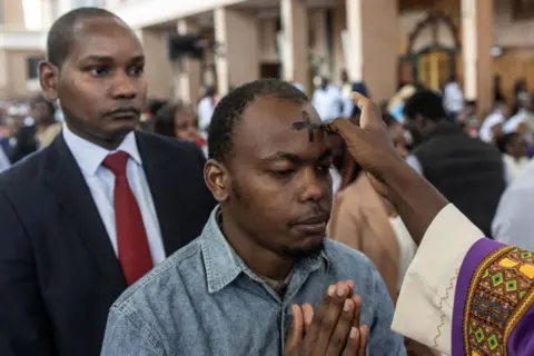 Simon Maina / AFP Seorang imam Katolik menerapkan abu pada dahi seorang pria di atas ketaatan Abu Rabu di Gereja Basilika Keluarga Suci.