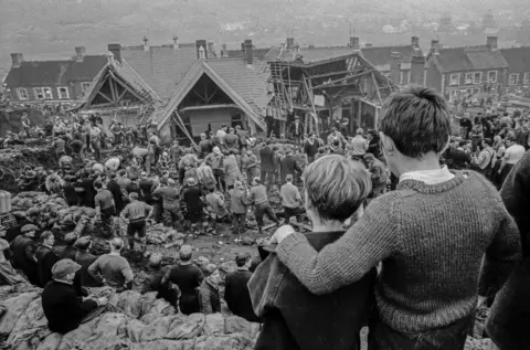 David Hurn A black and white photograph shot from behind of two young boys who look on at their destroyed school surrounded by scores of adults. The boys have their arms around one another.