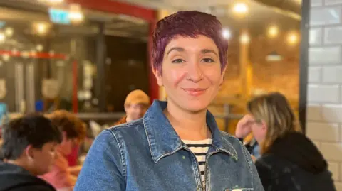Carla is standing near a white tiled wall in a café environment. She is wearing a denim top with a visible patch on the chest and striped sleeves. In the background, several people are seated at tables, and the setting includes exposed brick walls and overhead lighting.